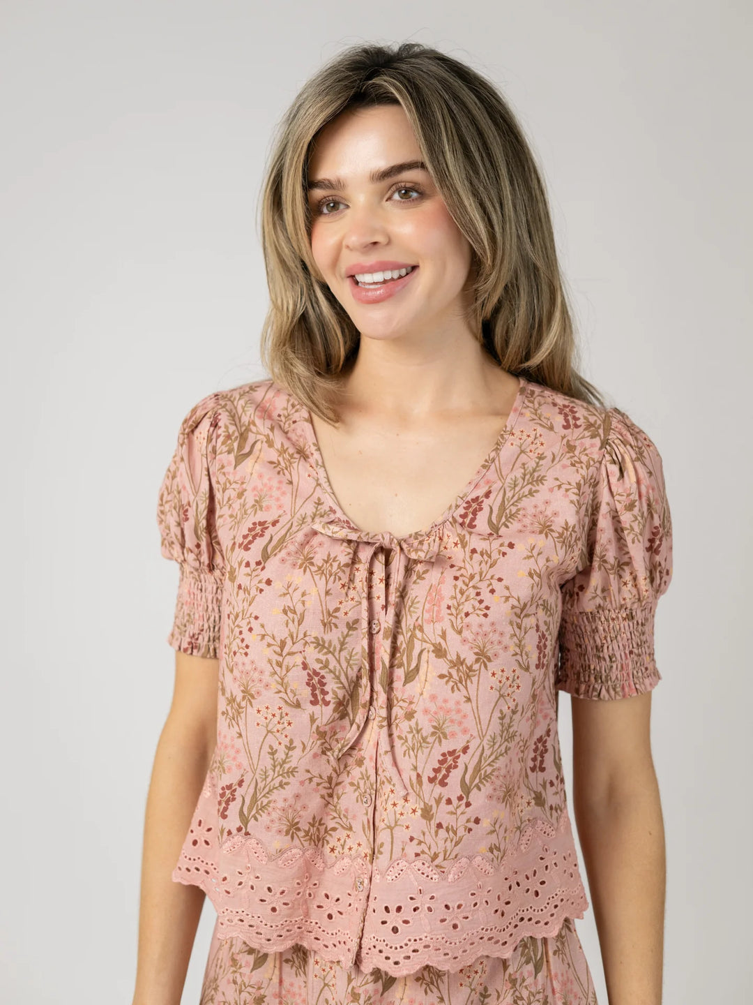 Front view of a woman smiling in the Louisa Top in Autumn Stems Rose, a pink floral cotton blouse with a scoop neckline, self-tie detail, puff sleeves, and smocked cuffs.