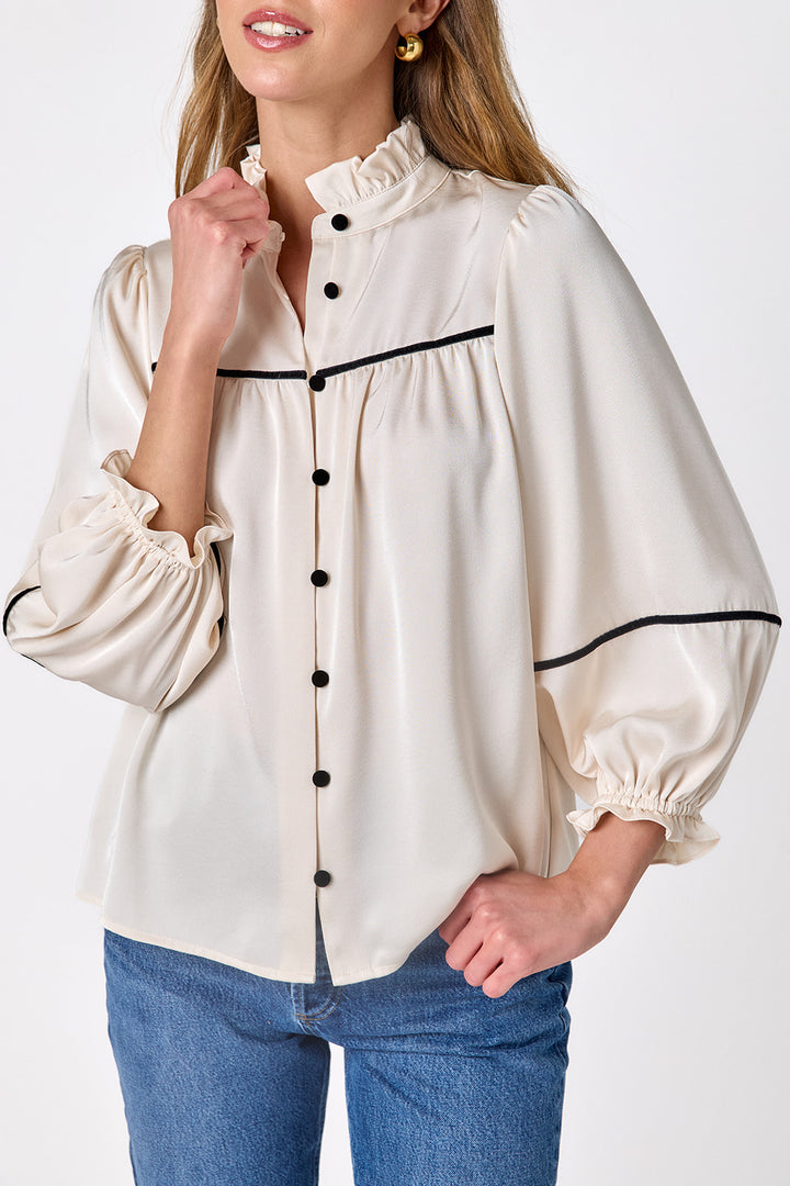 Close-up of the Crosby Worth Blouse in ivory highlighting the ruffled collar, black covered buttons, gathered yoke, contrast piping, and elasticized ruffle cuffs.