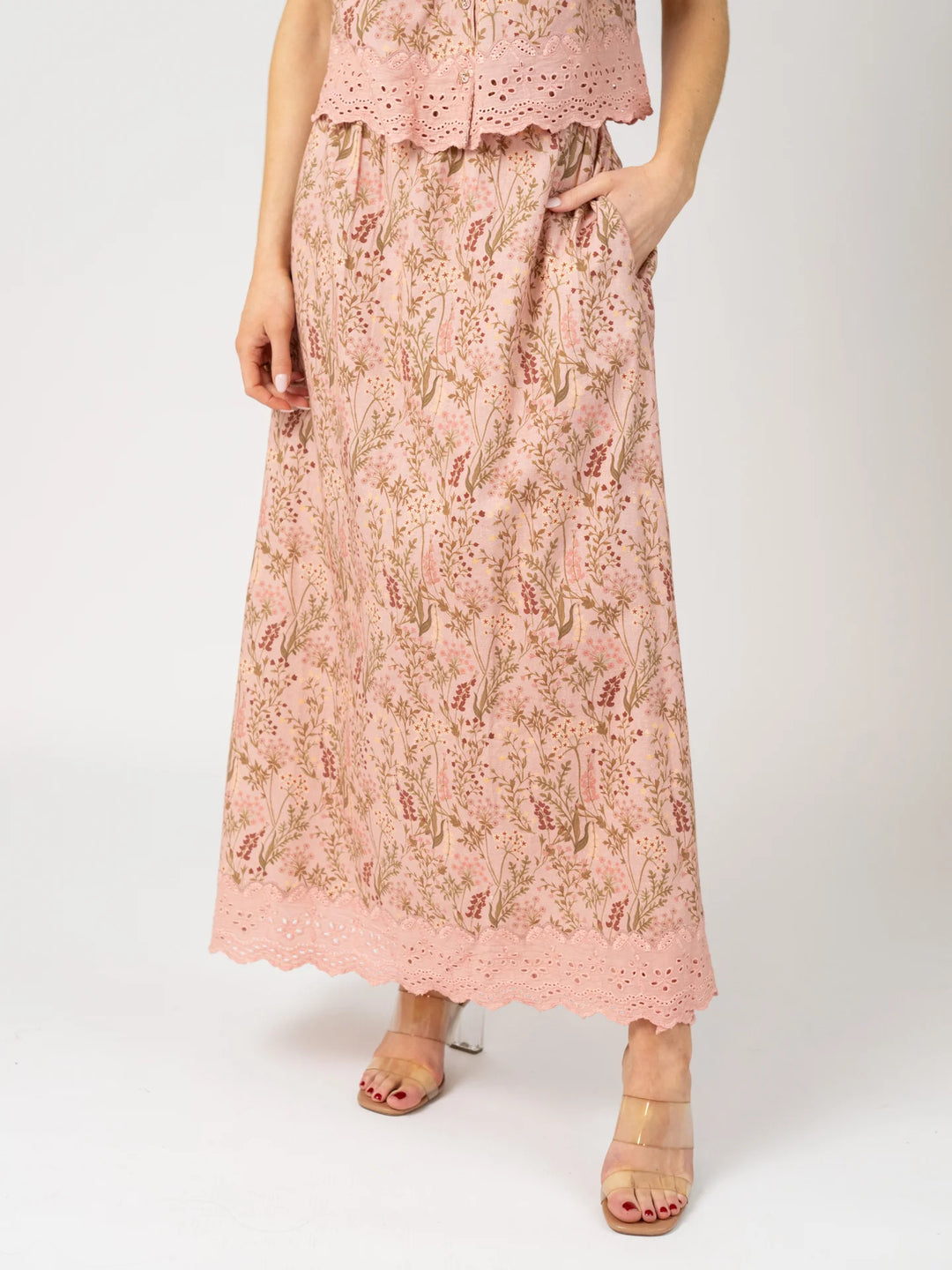 Close-up front view of a woman wearing the Markey Skirt in Autumn Stems Rose, an A-line pink floral cotton maxi skirt with a scalloped eyelet hem, hand tucked in the side pocket.