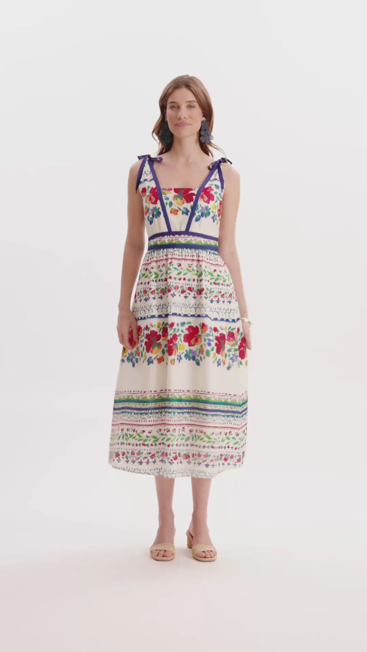 Full length view of Tyler Boe Chelsey Cotton Summer Soiree Dress in ivory with multicolor painterly floral print and navy grosgrain trim, shown front on-model, square V-neckline with shoulder-tie straps, fitted bodice, full midi skirt with bordered print bands, styled with raffia slides.