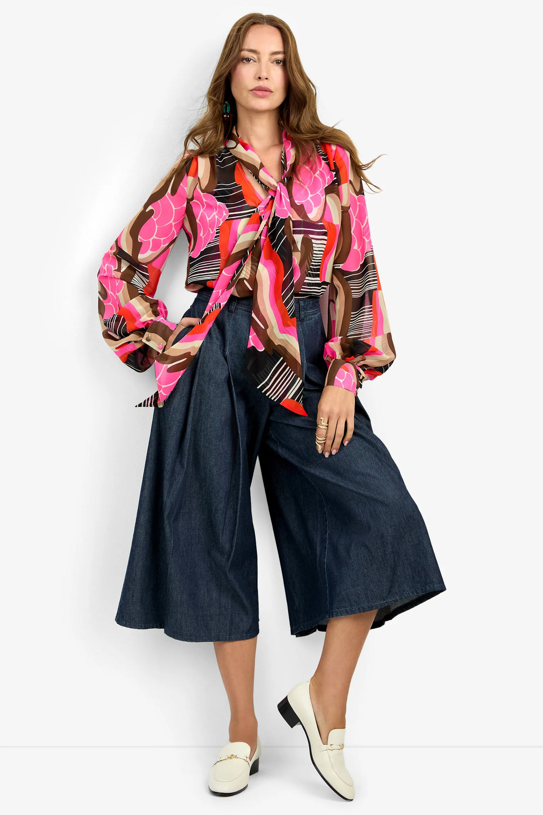 Trina Turk Pienza Top in pink, brown, and orange abstract print, front view, styled with dark denim culottes, statement neck tie visible.