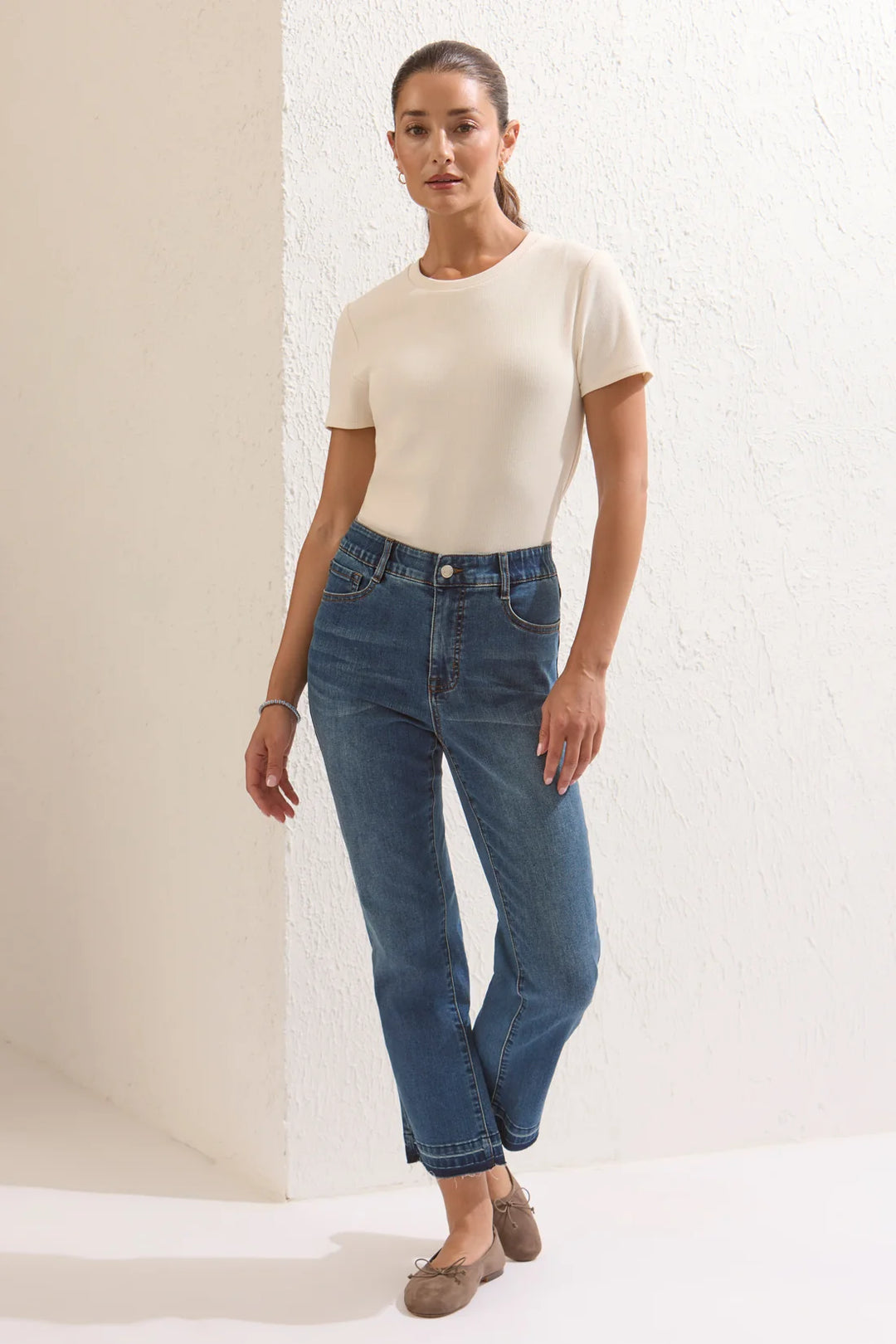 Full-body front view of a model wearing medium wash micro-flare jeans with a raw released hem, paired with a cream ribbed t-shirt and brown suede ballet flats.