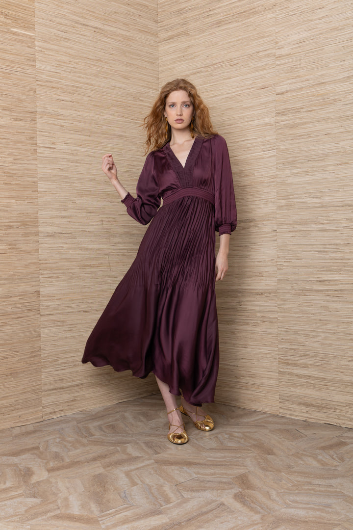 Hale Bob Yamileth V Neck Long Dress in Plum, full-length front view on model, showing pleat-trimmed V-neckline, gathered bodice, smocked waist, and flowing satin skirt, styled with gold drop earrings and gold flats.
