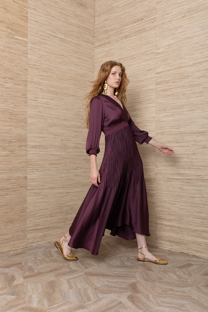Hale Bob Yamileth V Neck Long Dress in Plum, side view of model walking, showing pintuck pleating through the hip, asymmetric hem in motion, and blouson sleeves with smocked cuffs.