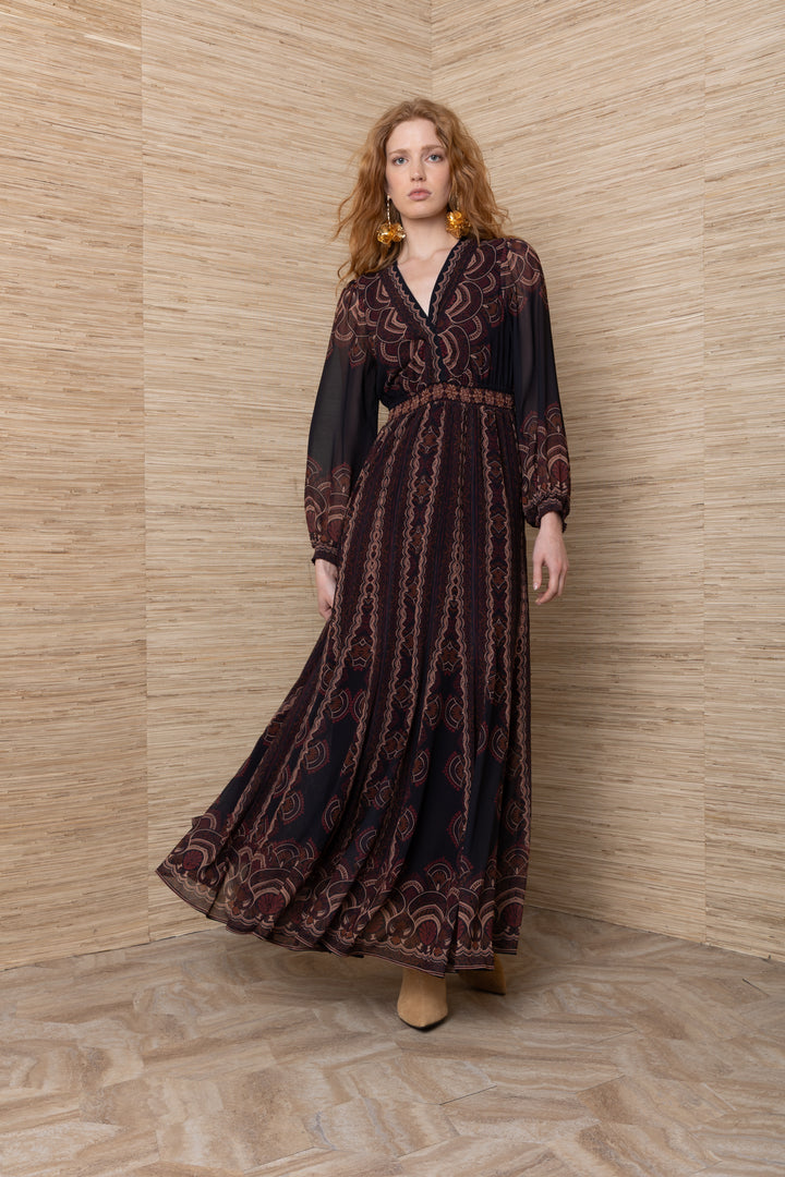 Hale Bob Iselda V Neck Maxi in Plum Multi, full-length front view on model, showing crossover V-neckline, smocked waist, sheer blouson sleeves, and full maxi skirt with engineered border print, styled with gold floral earrings and tan suede boots.