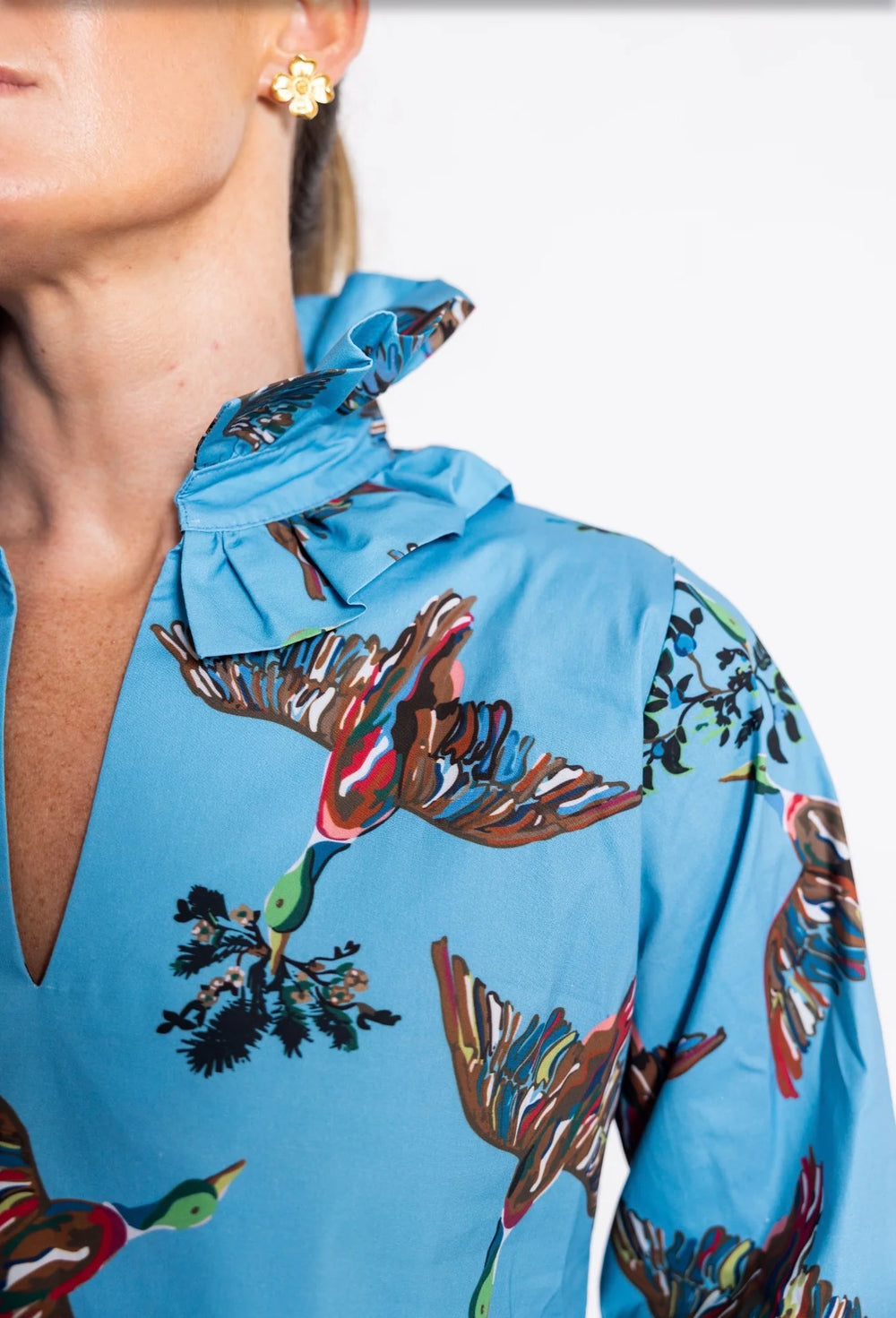 Close up: House of MBR Harper Top in Berry Grove© print, showing tunic-length silhouette in blue with illustrated bird print, double ruffle mandarin collar, and push-up sleeves, styled with dark denim in an ornate interior.