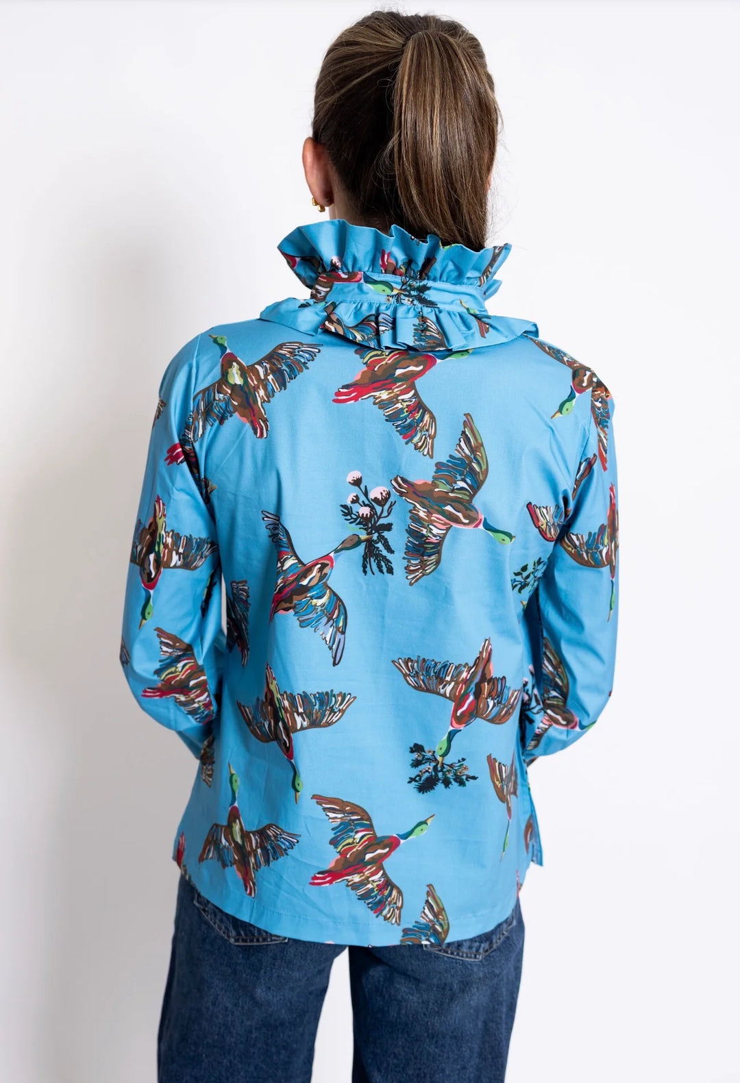 Back view: House of MBR Harper Top in Berry Grove© print, showing tunic-length silhouette in blue with illustrated bird print, double ruffle mandarin collar, and push-up sleeves, styled with dark denim in an ornate interior.