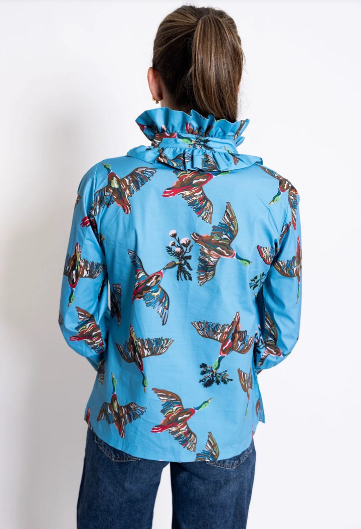 Back view: House of MBR Harper Top in Berry Grove© print, showing tunic-length silhouette in blue with illustrated bird print, double ruffle mandarin collar, and push-up sleeves, styled with dark denim in an ornate interior.