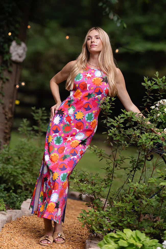 Alden Adair Berkley Maxi Dress in hot pink with multicolor rainbow starburst print, shown front on-model, sleeveless crew neckline, column maxi silhouette with side seam pockets, styled with woven slides taken in a garden.