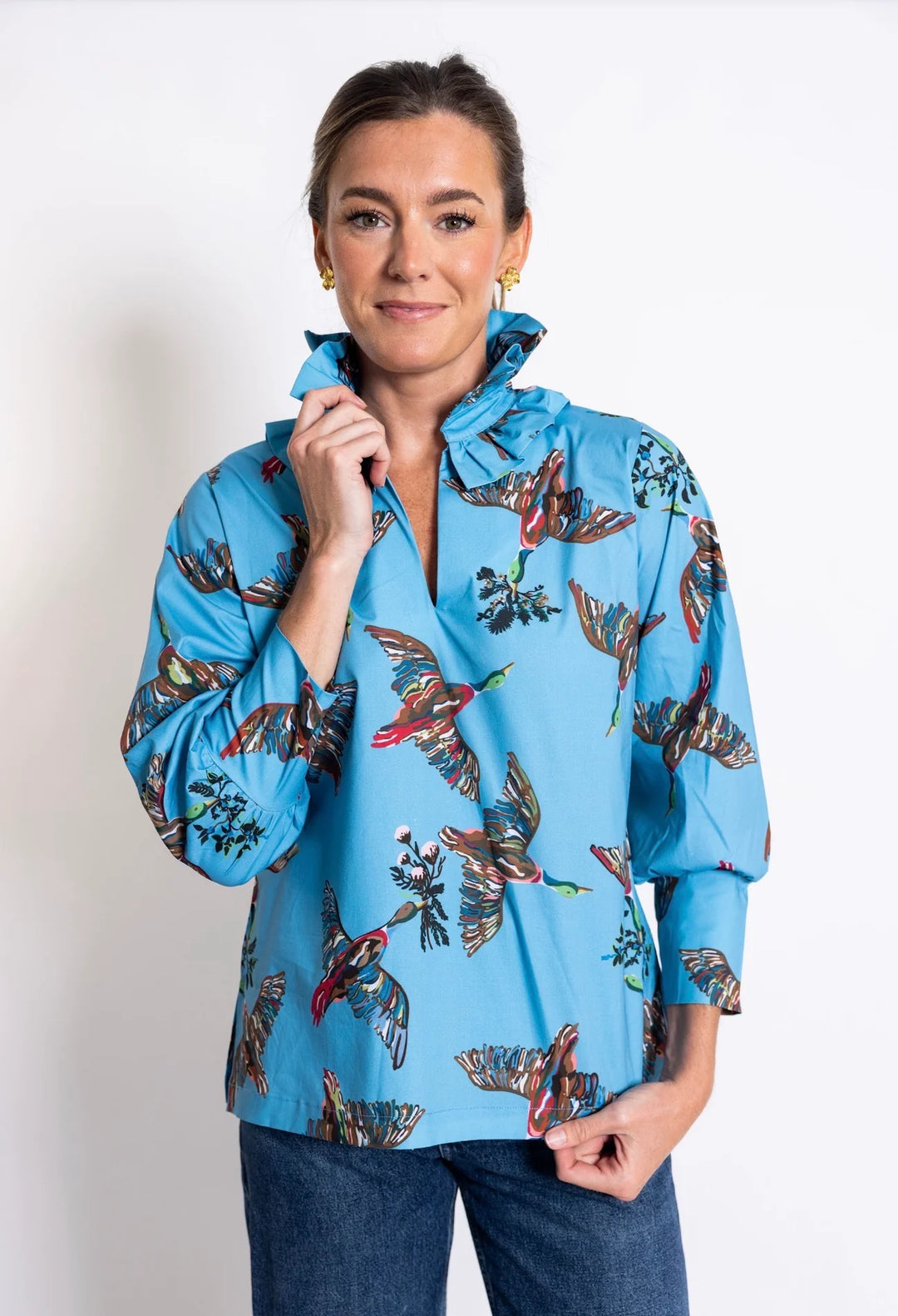 Front view - House of MBR Harper Top in Berry Grove© print, showing tunic-length silhouette in blue with illustrated bird print, double ruffle mandarin collar, and push-up sleeves, styled with dark denim in an ornate interior.