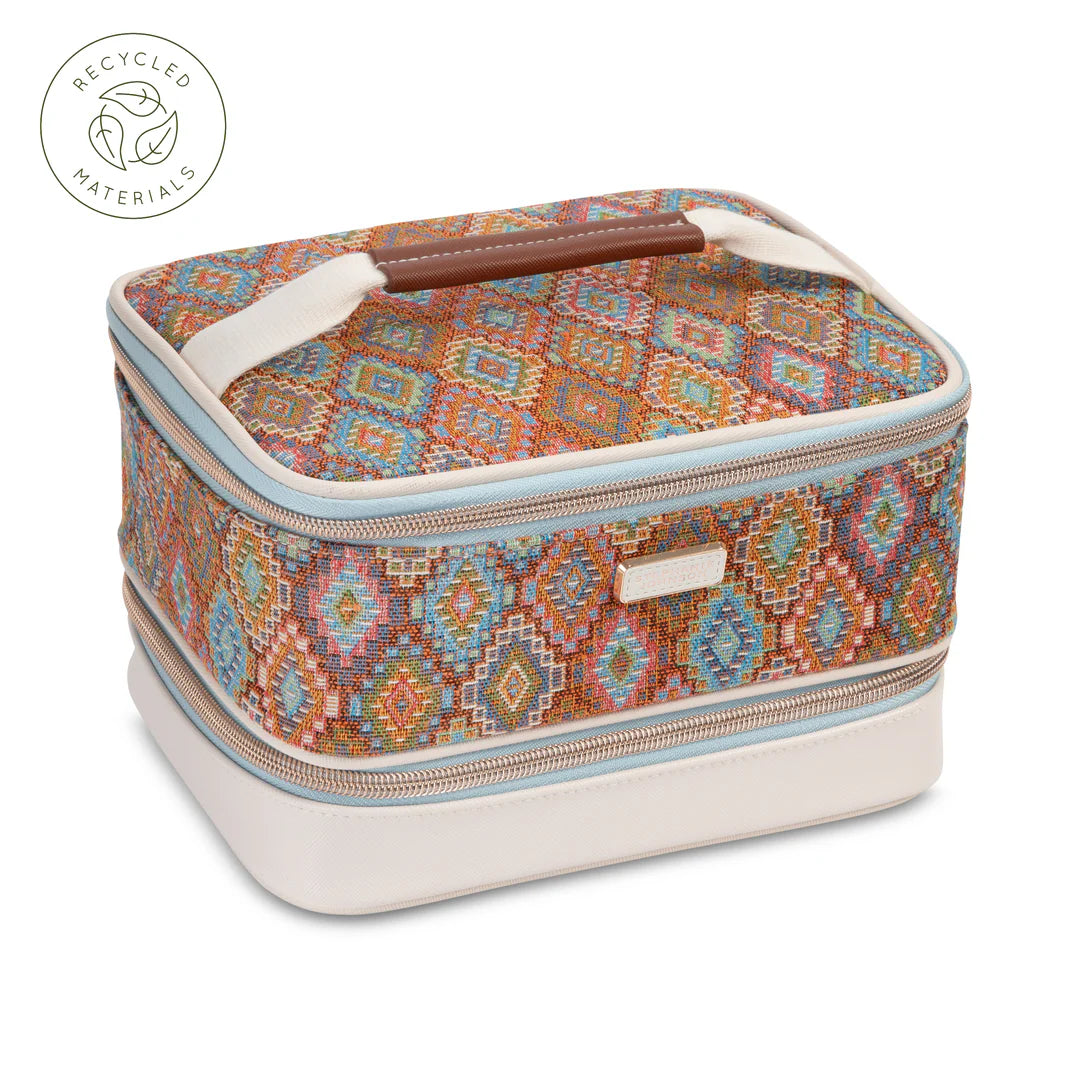 Front view: Stephanie Johnson Jenny Train Case in Bodrum Kilim print, showing woven diamond-pattern exterior in rust, blue, and coral tones, ivory faux-leather base trim, and rolled brown leather top handle.