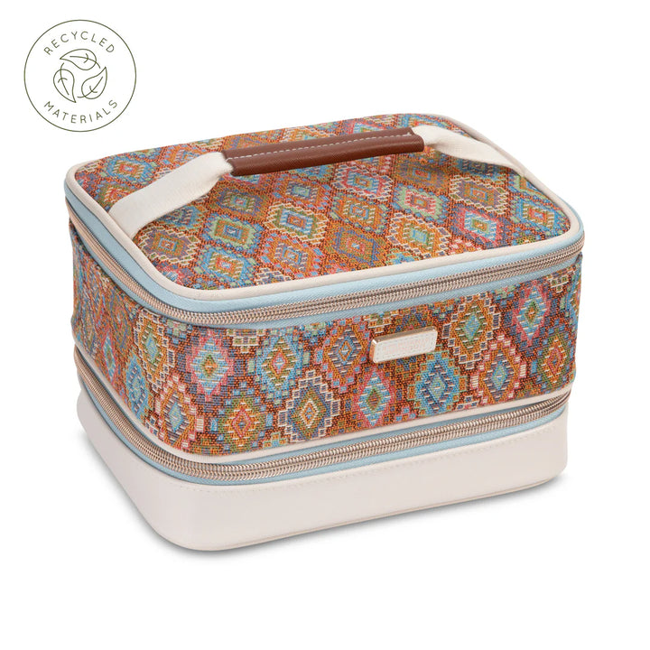 Front view: Stephanie Johnson Jenny Train Case in Bodrum Kilim print, showing woven diamond-pattern exterior in rust, blue, and coral tones, ivory faux-leather base trim, and rolled brown leather top handle.