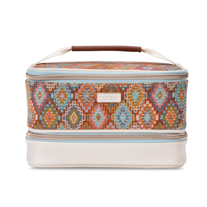 Front full view:  Stephanie Johnson Jenny Train Case in Bodrum Kilim print, showing woven diamond-pattern exterior in rust, blue, and coral tones, ivory faux-leather base trim, and rolled brown leather top handle.  