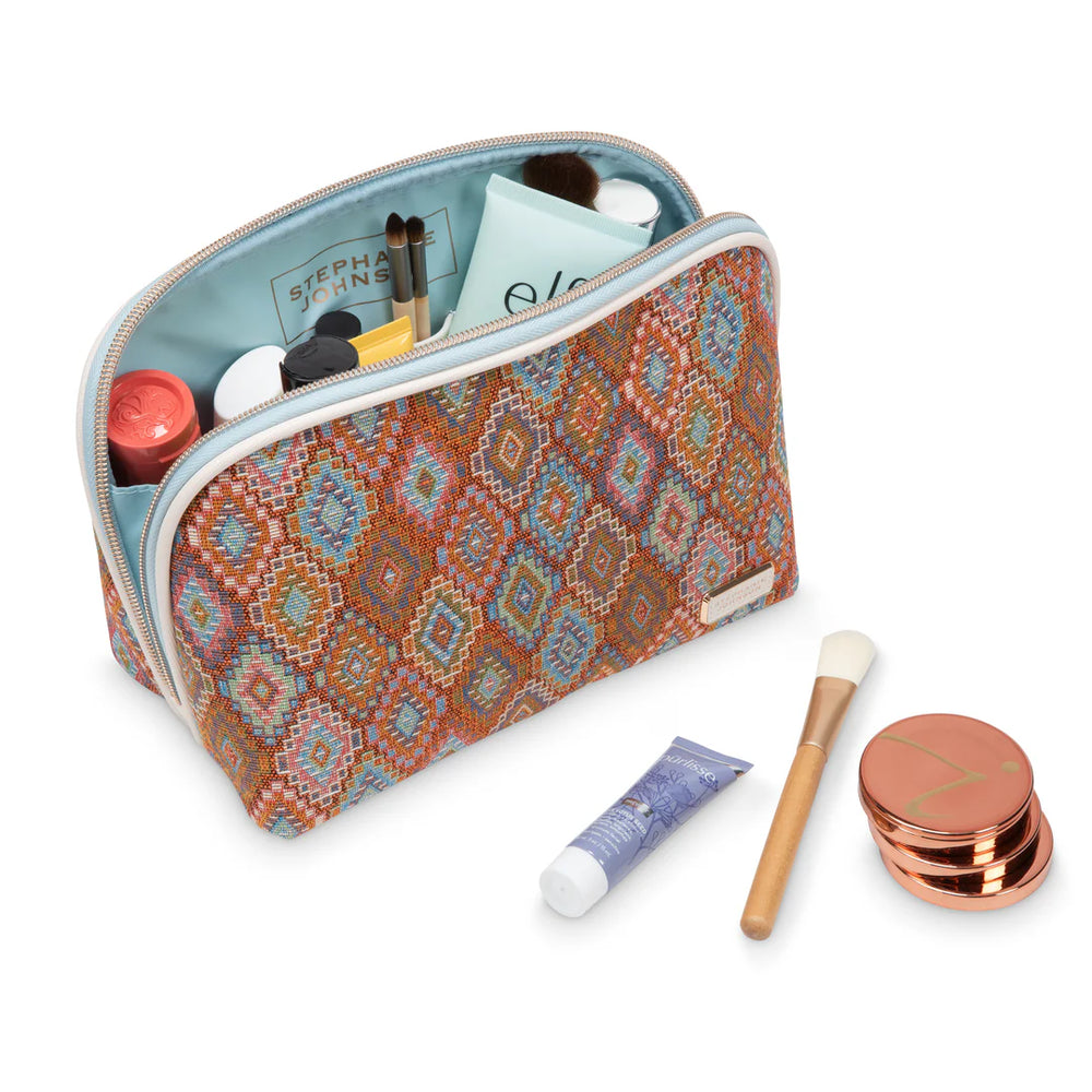 Open/styled view: Stephanie Johnson Lola Makeup Bag open and filled with skincare and makeup products, showing pale blue interior lining and elastic stash pockets, with makeup brush and compact styled alongside.