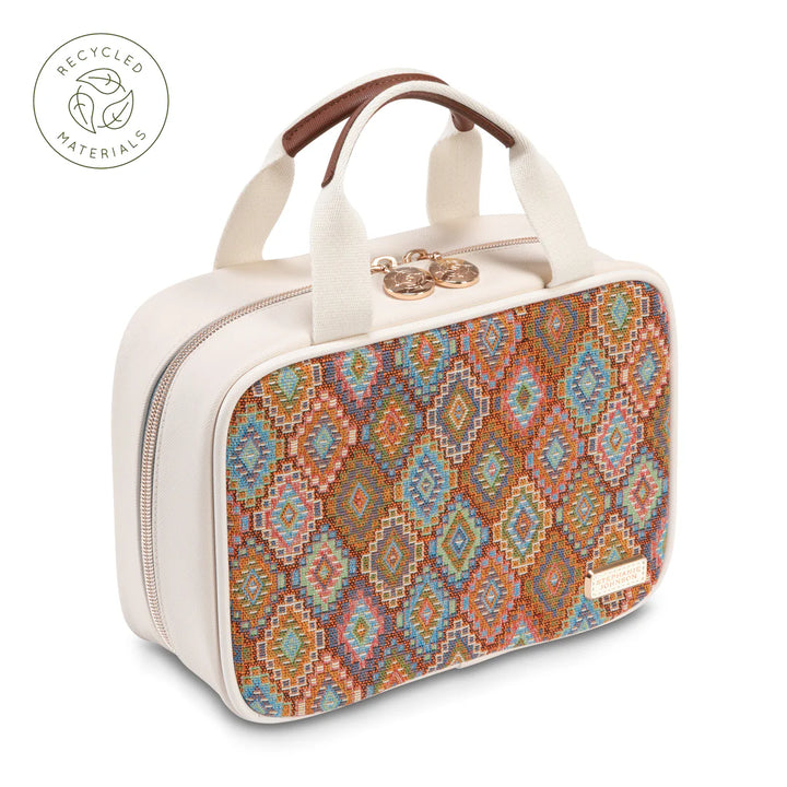 Angled top-down view: Stephanie Johnson ML Traveler in Bodrum Kilim print, shown diamond-tilted to highlight woven diamond-pattern panel in rust, blue, and coral against ivory faux-leather body, with canvas-and-leather top handle.