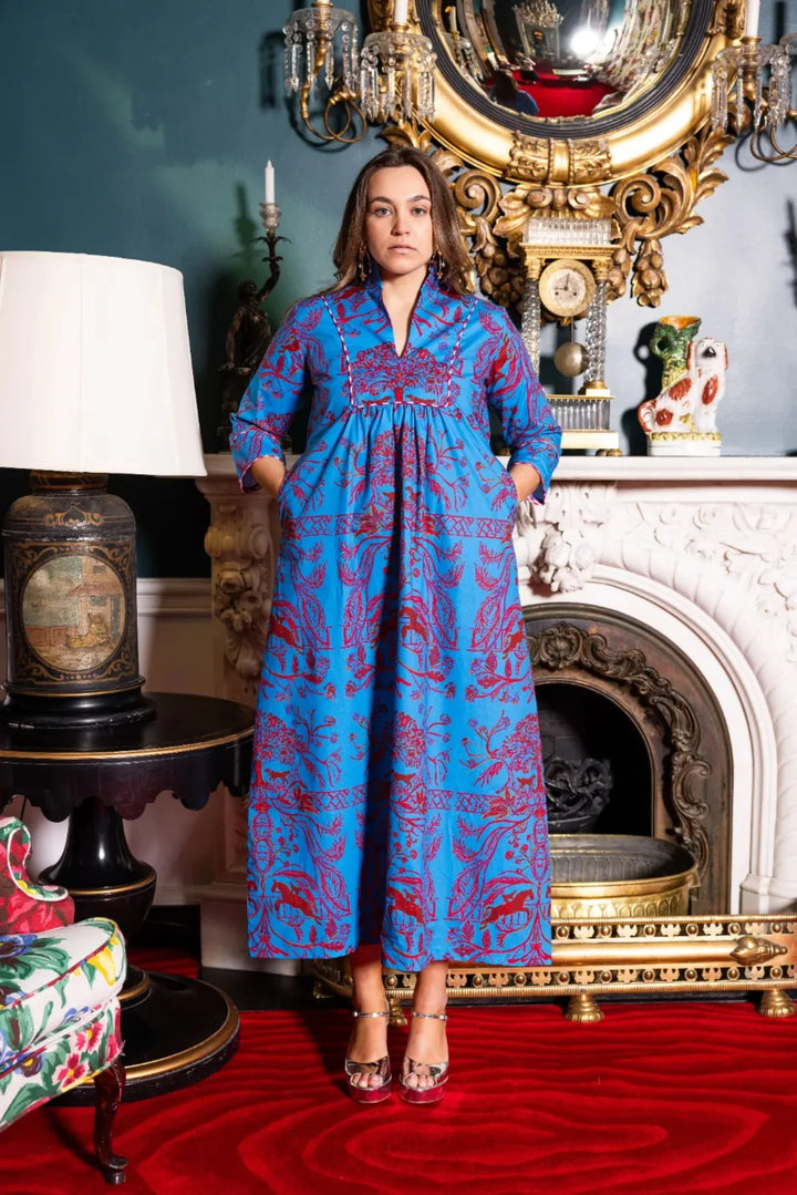 Front view, styled lifestyle: House of MBR Deb Dress Long in Bit and Boll© print, showing full-length caftan silhouette with vivid blue ground and red equestrian-inspired print, banded collar, empire seam, and 3/4 fluted sleeves, styled in an ornate interior with hands in pockets.