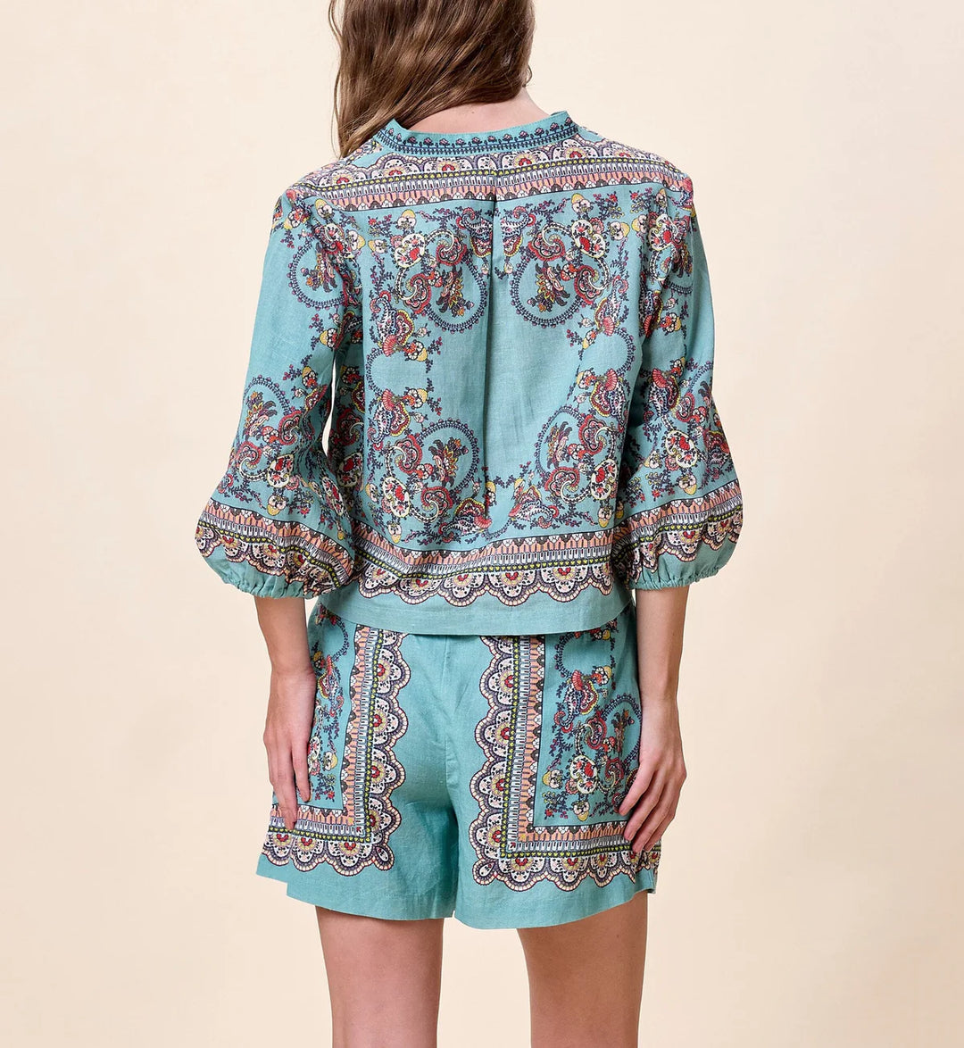 Model wearing Cleobella Noelle Blouse in teal paisley print, cropped relaxed fit with button-front closure and band collar, styled with matching printed shorts.