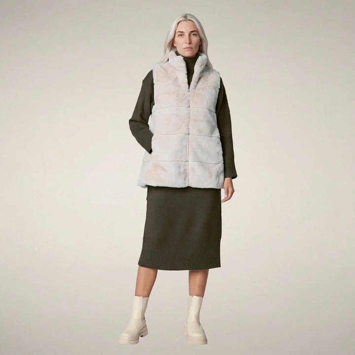 Front view of a woman wearing the Nives Faux Fur Reversible Vest in shore beige over an olive knit sweater dress and cream boots.