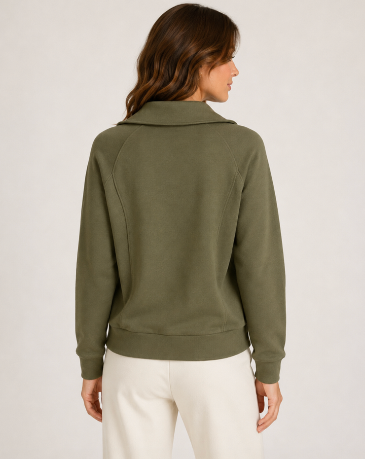 A woman shown from the back wearing an olive green long-sleeve top with a mock neck and white pants, photographed against a plain light gray studio background.