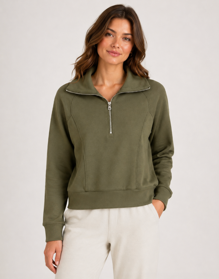 A woman wearing an olive green half-zip sweatshirt with a stand collar and silver zipper, paired with off-white sweatpants, photographed against a light gray studio background.