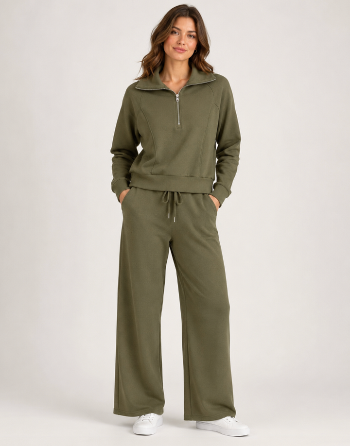 A woman modeling an olive green two-piece loungewear set consisting of a half-zip pullover and matching wide-leg sweatpants, paired with white sneakers, against a light gray studio background.