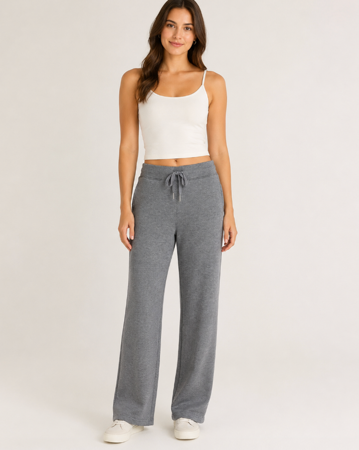 A woman modeling a pair of heather gray wide-leg sweatpants with a drawstring waist, paired with a white ribbed camisole and white sneakers, against a plain light gray studio background.