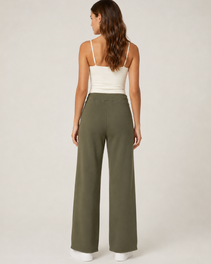 A woman shown from the back wearing olive green wide-leg pants and a white spaghetti-strap camisole top, standing against a plain light gray studio background.