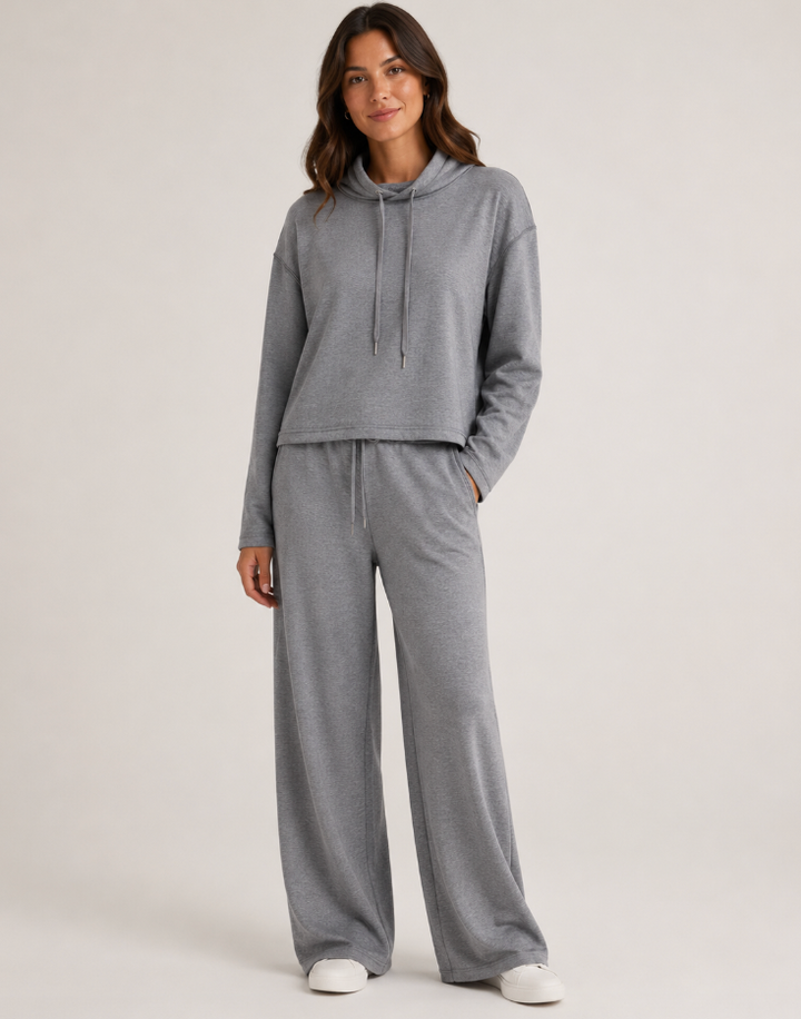 A woman modeling a matching gray lounge set consisting of a cropped long-sleeve hoodie with drawstrings and wide-leg sweatpants, photographed against a plain light gray studio background.