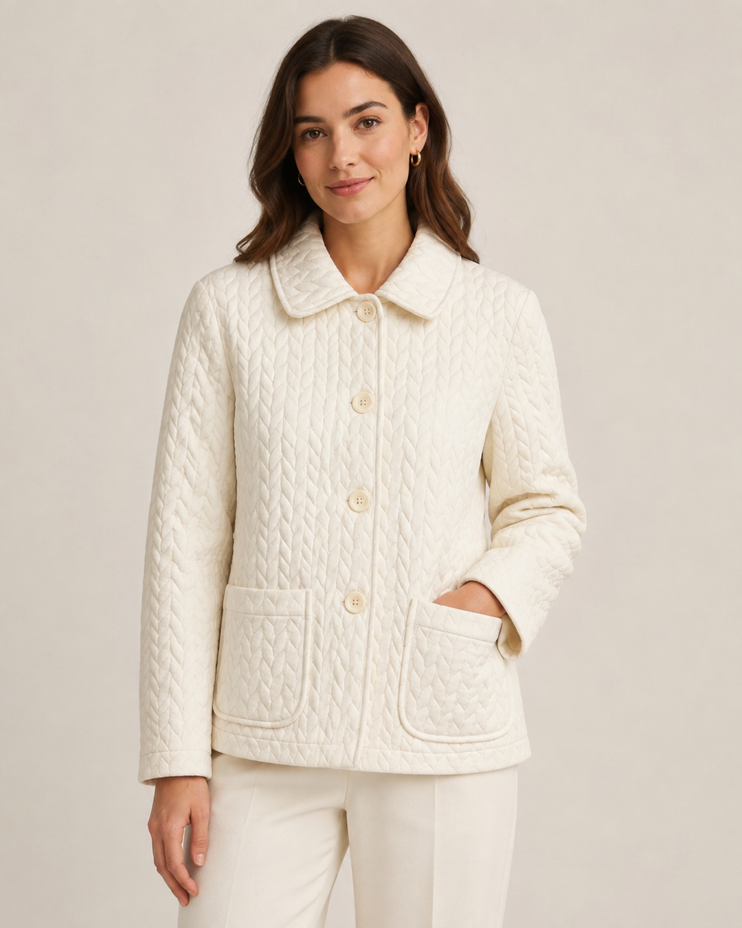 A woman wearing a cream-colored quilted cable-knit jacket with a collar and front buttons, paired with matching cream pants, against a solid light beige background.