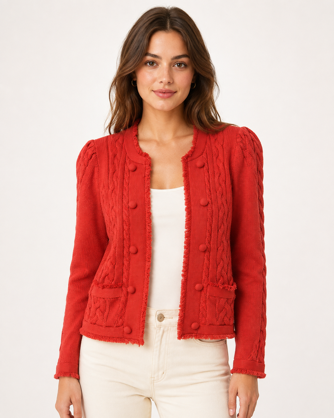 A woman wearing a bright red cable-knit cropped jacket with decorative buttons and frayed trim, styled over a white top and cream pants against a plain white studio background.