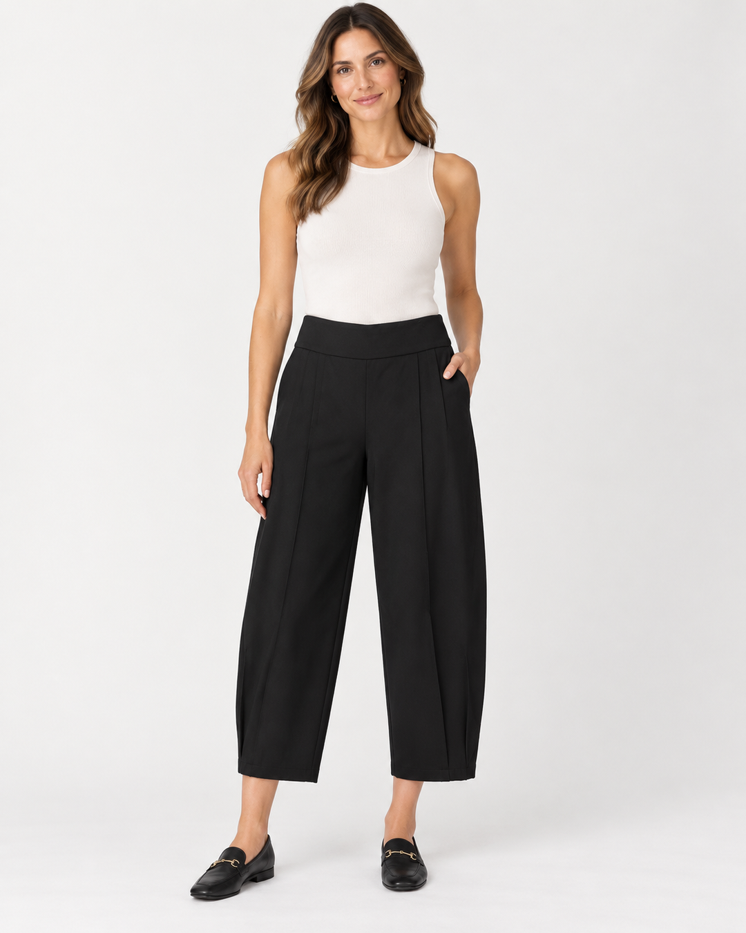 Front view of a woman with long wavy brown hair wearing a white ribbed tank top and Elliott Lauren black barrel-leg cropped pants, one hand in pocket, paired with black loafers, against a light gray background.