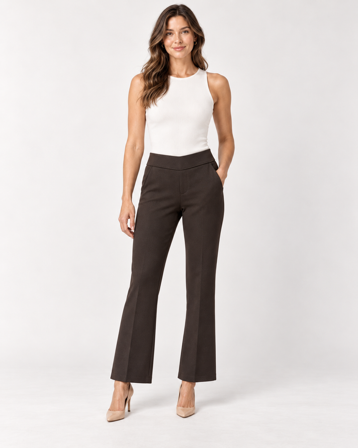 Front view of a woman with long wavy brown hair wearing a white ribbed tank top and Elliott Lauren chocolate brown baby boot pants with a comfort waistband, one hand in pocket, paired with nude pointed pumps, against a light gray background.
