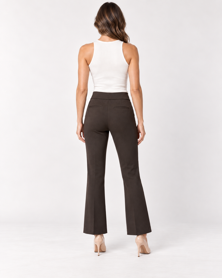 Back view of a woman with long wavy brown hair wearing a white ribbed tank top and Elliott Lauren chocolate brown baby boot pants with back besom pockets, paired with nude pointed pumps, against a light gray background.