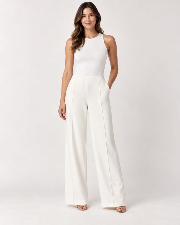 Front view of a woman with long wavy brown hair wearing a white ribbed tank top and Elliott Lauren wide-leg ponte pants in winter white with a front seam crease, one hand in pocket, paired with nude open-toe heels, against a light gray background.
