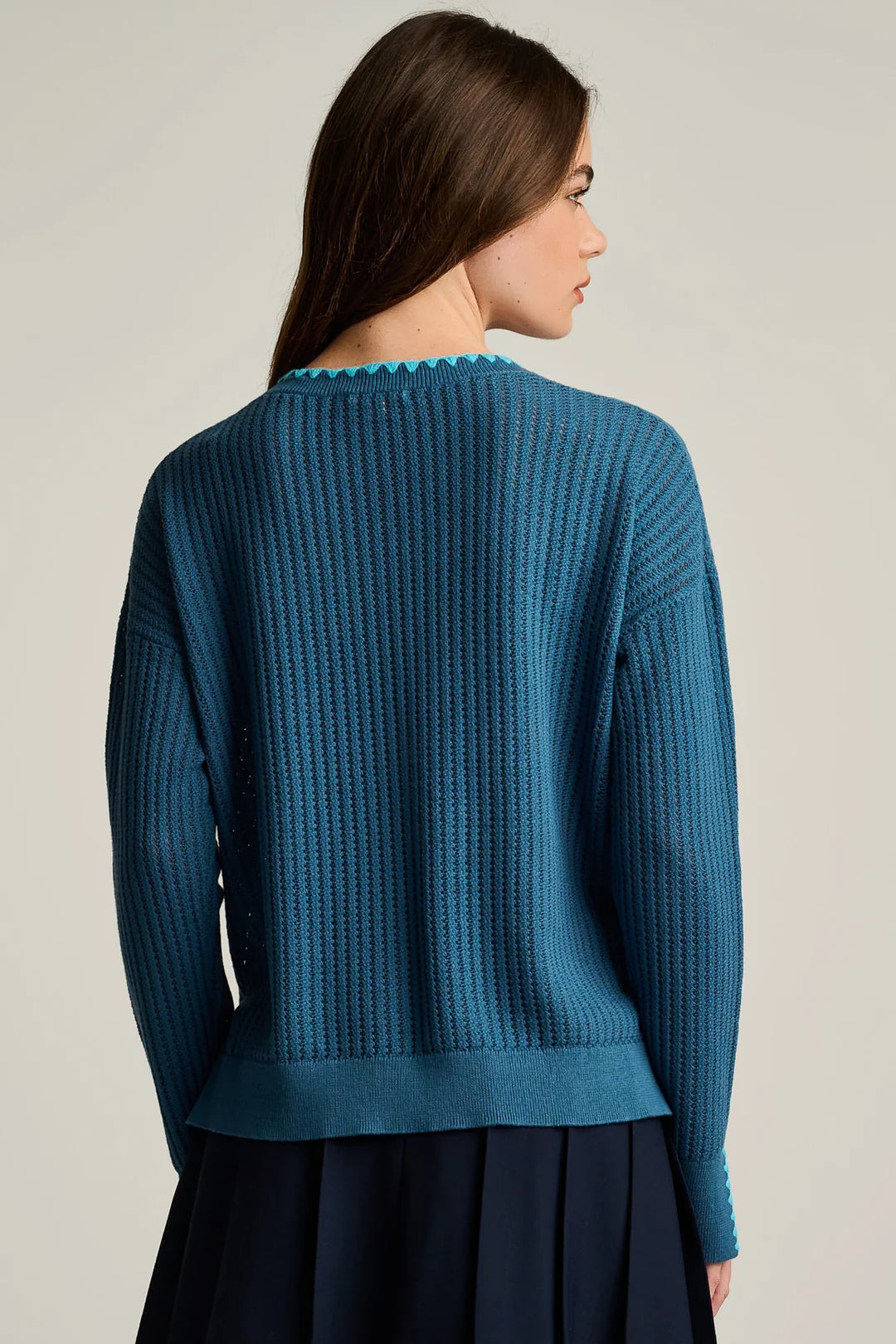 A woman shown from the back wearing a teal blue ribbed knit sweater with a scalloped neckline, paired with a dark navy skirt, against a plain light gray studio background.