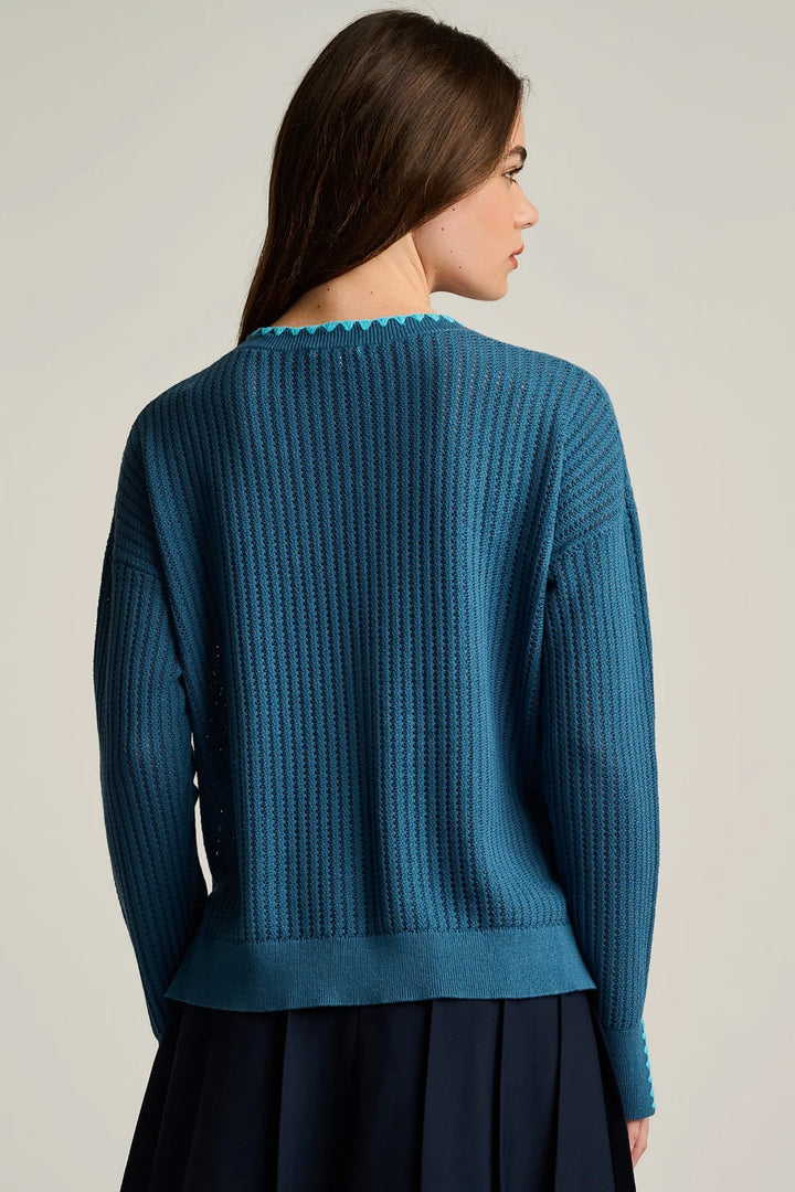 A woman shown from the back wearing a teal blue ribbed knit sweater with a scalloped neckline, paired with a dark navy skirt, against a plain light gray studio background.