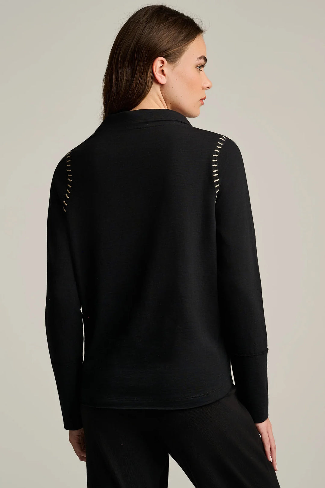 A woman shown from the back wearing a black long-sleeve top with decorative metallic stud detailing along the shoulder seams, against a plain light gray studio background.