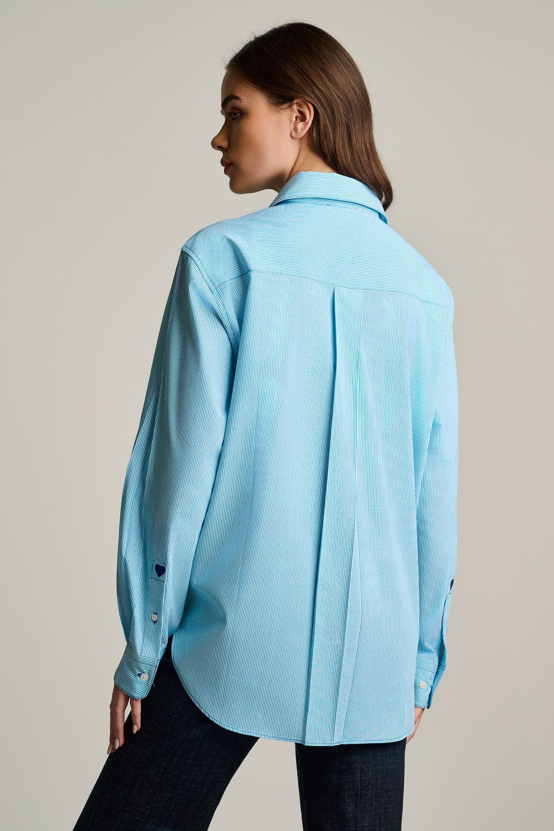 A woman shown from the back wearing a light blue long-sleeve button-up shirt and dark jeans, photographed against a plain light beige studio background.