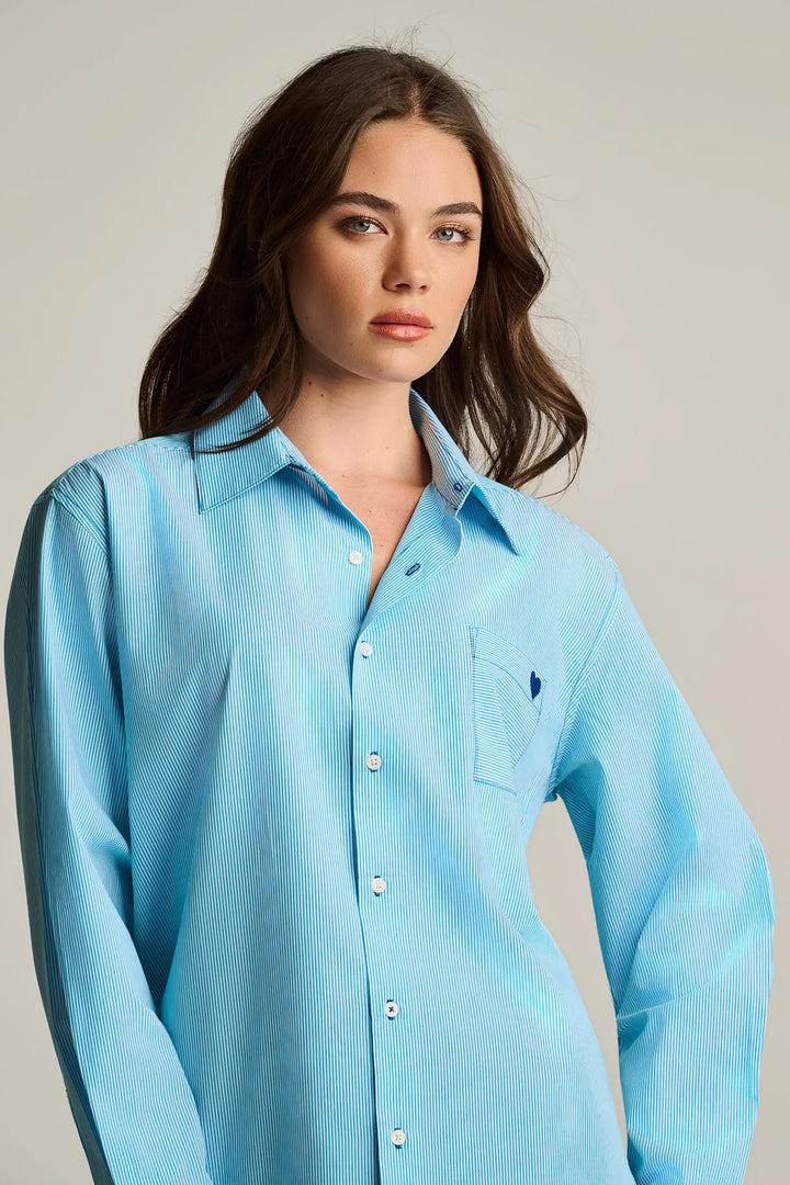 A woman wearing a light blue long-sleeve button-up shirt with a chest pocket and a small embroidered logo, photographed against a plain light gray studio background.