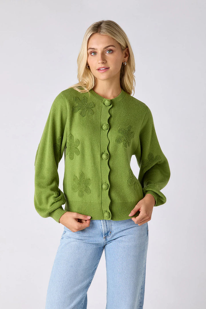Model wearing Crosby Colson Cardigan in grass, front view, styled with light-wash straight-leg jeans, showing tonal daisy jacquard pattern and crochet-covered button closure, on grey background.