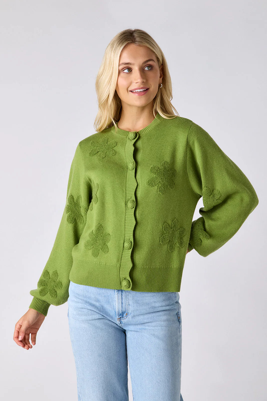 Model wearing Crosby Colson Cardigan in grass, front view, styled with light-wash straight-leg jeans, showing tonal daisy jacquard pattern and crochet-covered button closure, on grey background.