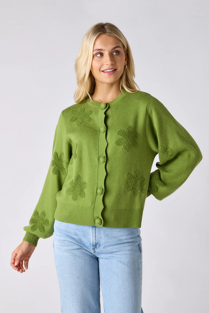 Model wearing Crosby Colson Cardigan in grass, front view, styled with light-wash straight-leg jeans, showing tonal daisy jacquard pattern and crochet-covered button closure, on grey background.