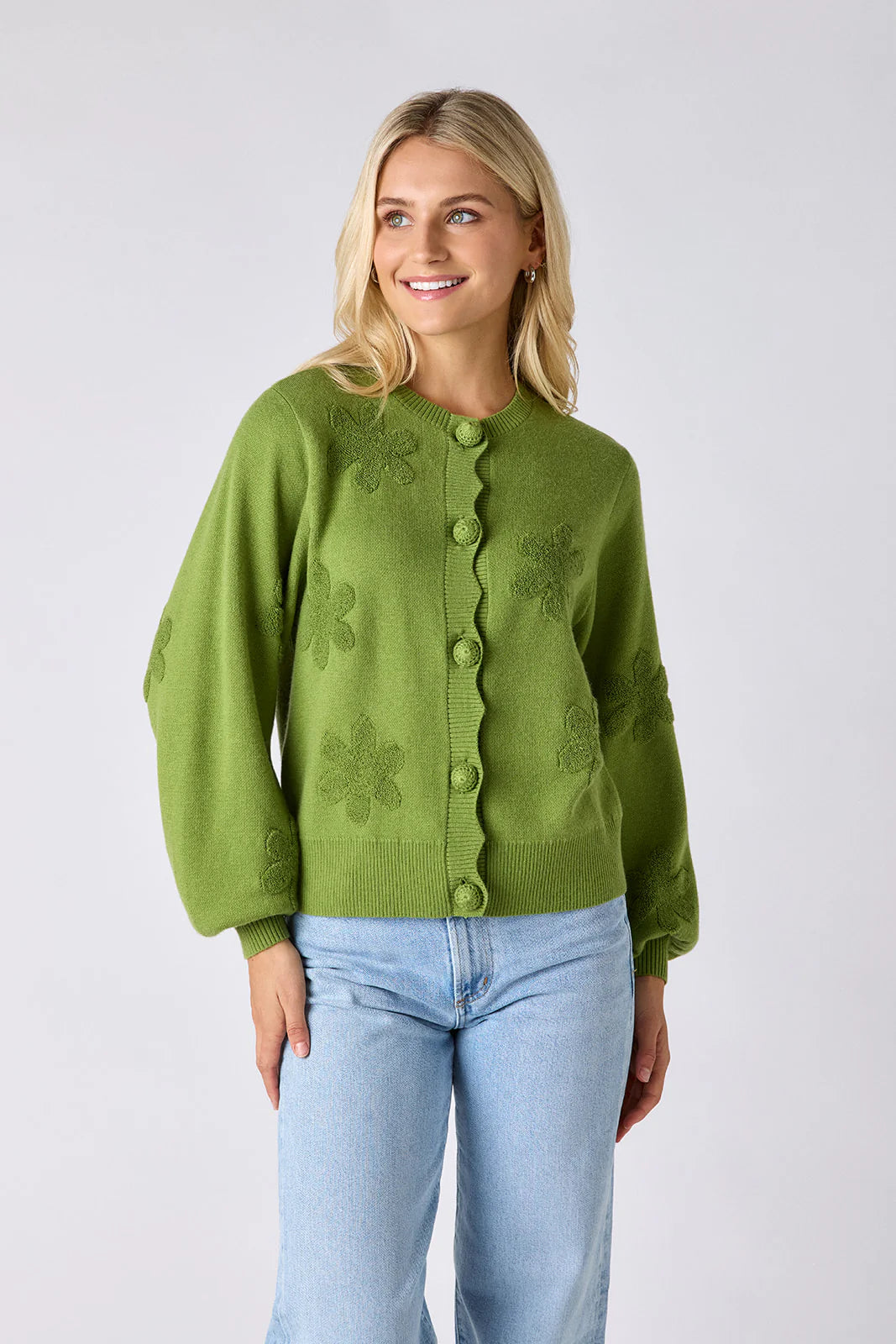 Model wearing Crosby Colson Cardigan in grass, front view, styled with light-wash straight-leg jeans, showing tonal daisy jacquard pattern and crochet-covered button closure, on grey background.