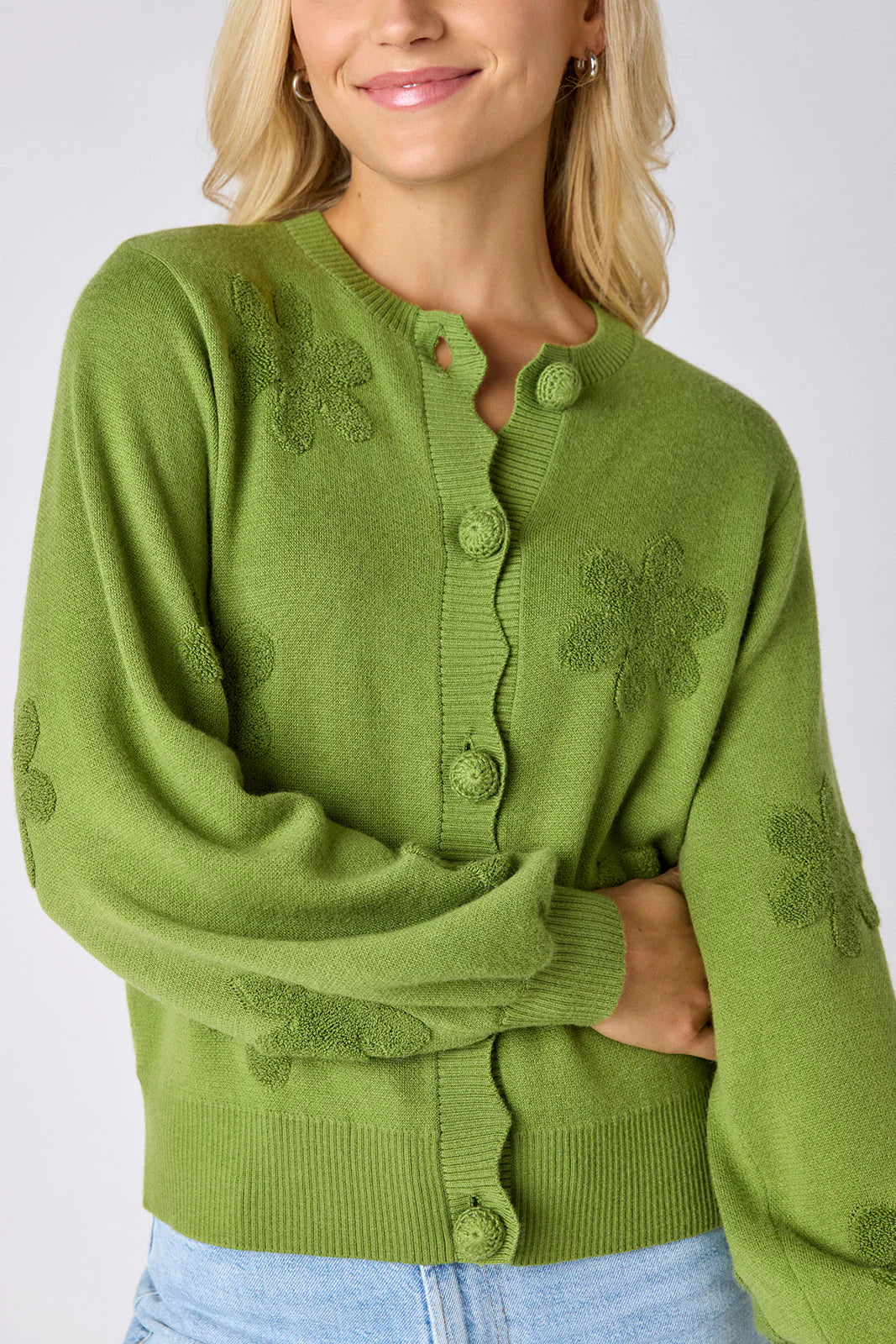 Model wearing Crosby Colson Cardigan in grass, front view, styled with light-wash straight-leg jeans, showing tonal daisy jacquard pattern and crochet-covered button closure, on grey background.
