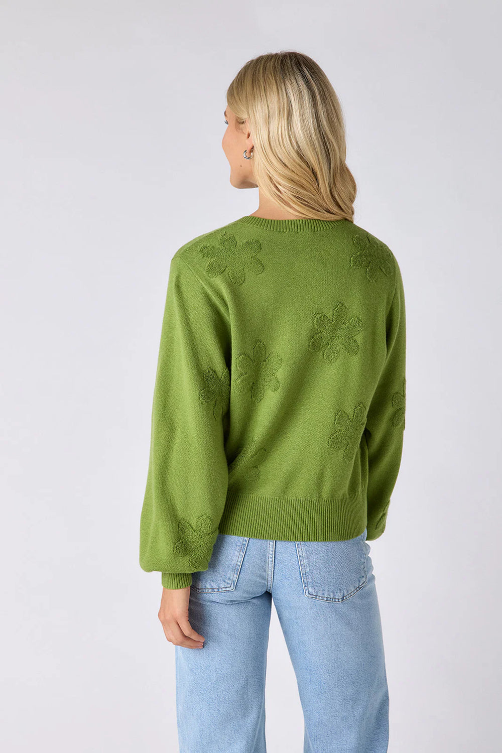 Model wearing Crosby Colson Cardigan in grass, front view, styled with light-wash straight-leg jeans, showing tonal daisy jacquard pattern and crochet-covered button closure, on grey background. - back