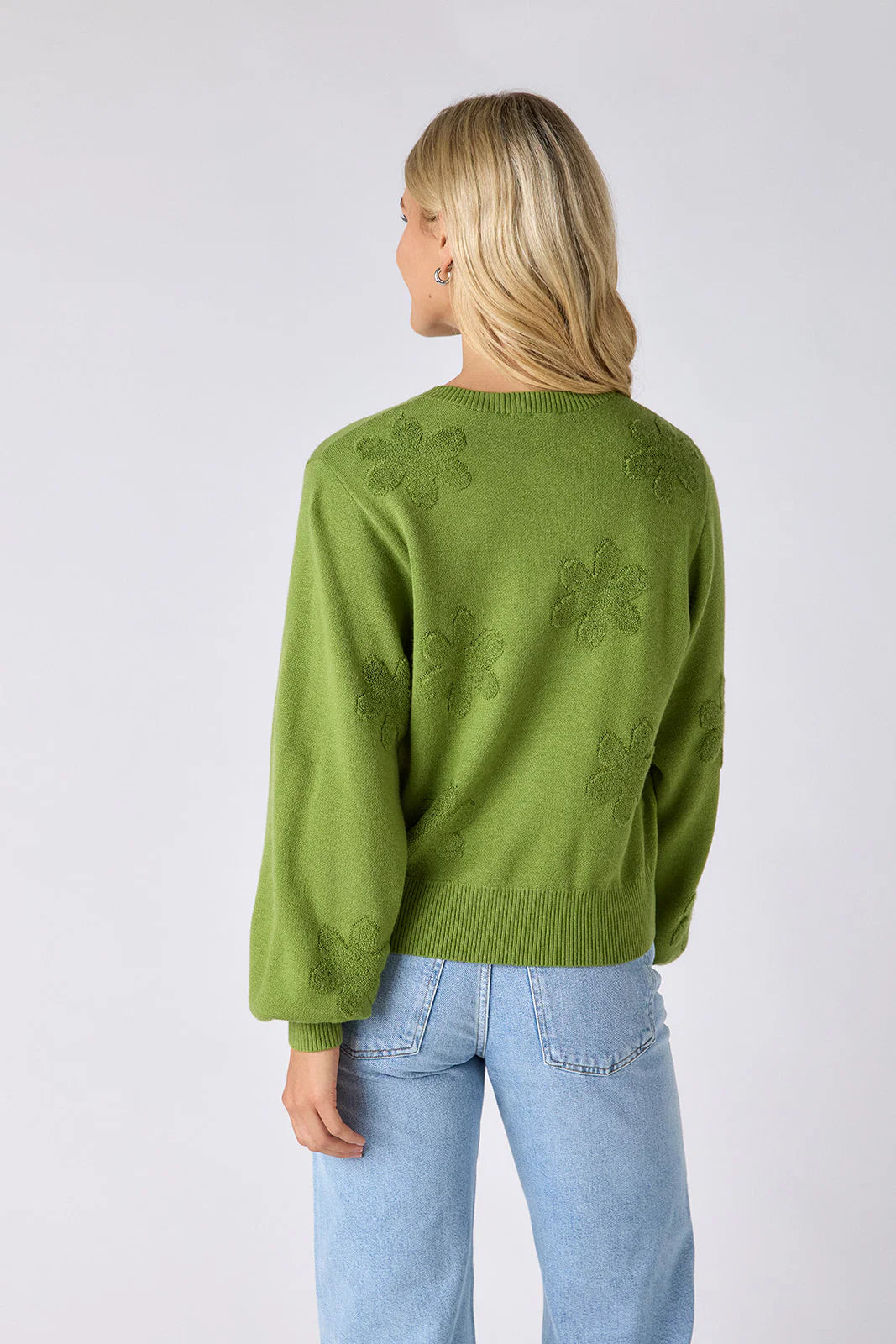Model wearing Crosby Colson Cardigan in grass, front view, styled with light-wash straight-leg jeans, showing tonal daisy jacquard pattern and crochet-covered button closure, on grey background. - back