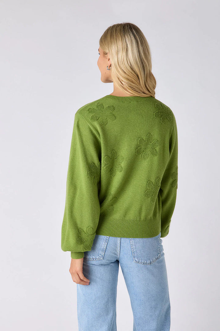 Model wearing Crosby Colson Cardigan in grass, front view, styled with light-wash straight-leg jeans, showing tonal daisy jacquard pattern and crochet-covered button closure, on grey background. - back