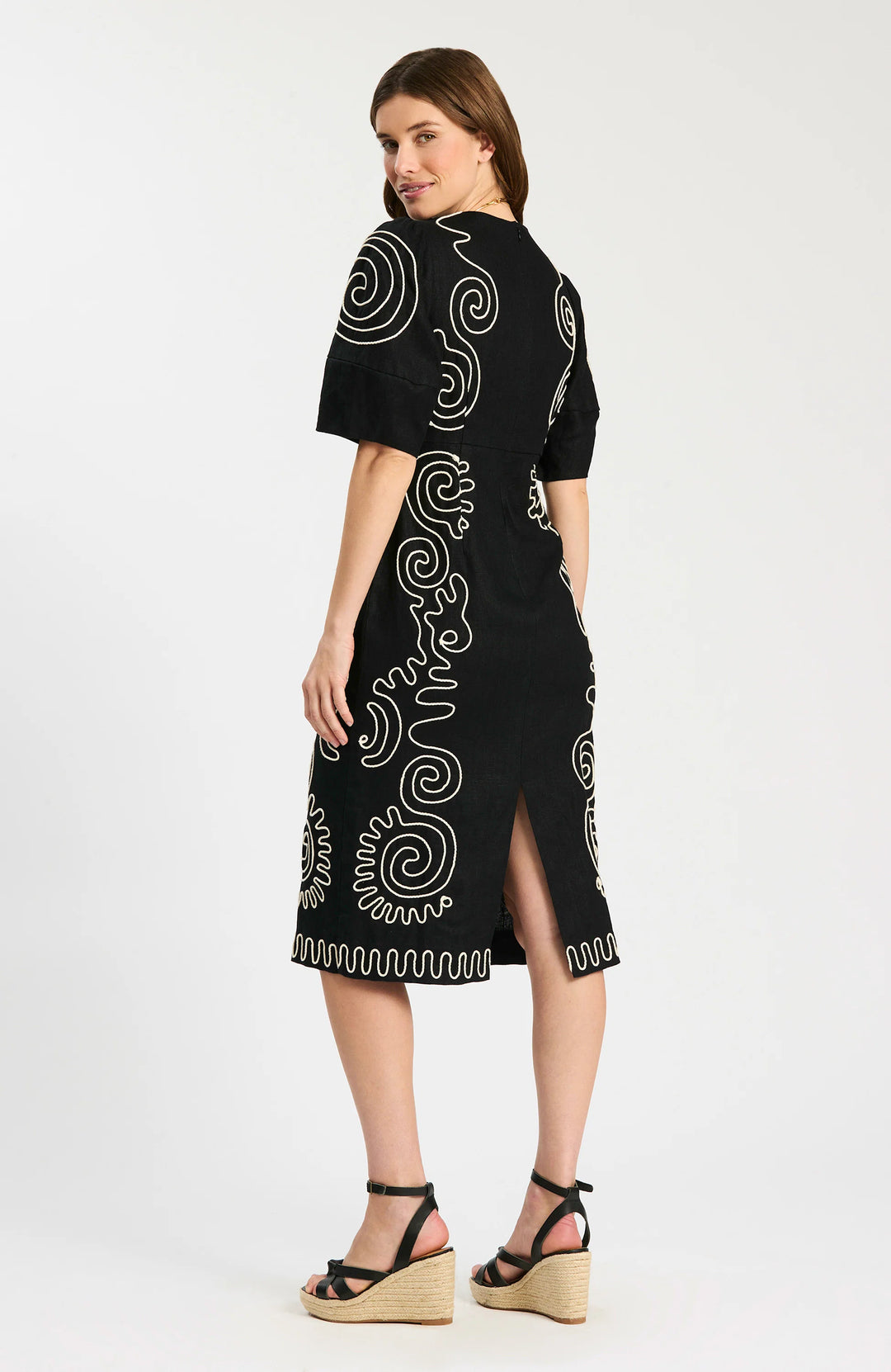 Model wearing Tyler Boe Posey Linen Embroidered Midi Dress in black with cream spiral cord embroidery, styled with black espadrille wedge sandals and gold necklace, full-length front view on white background. - back