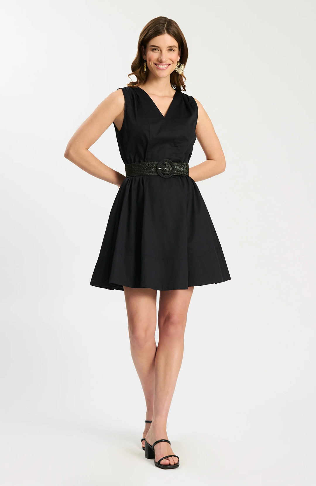 Model wearing Tyler Boe Emory Sleeveless Wrap Dress in black, front view belted with woven rattan belt, styled with black block-heel sandals and gold earrings, on white background.
