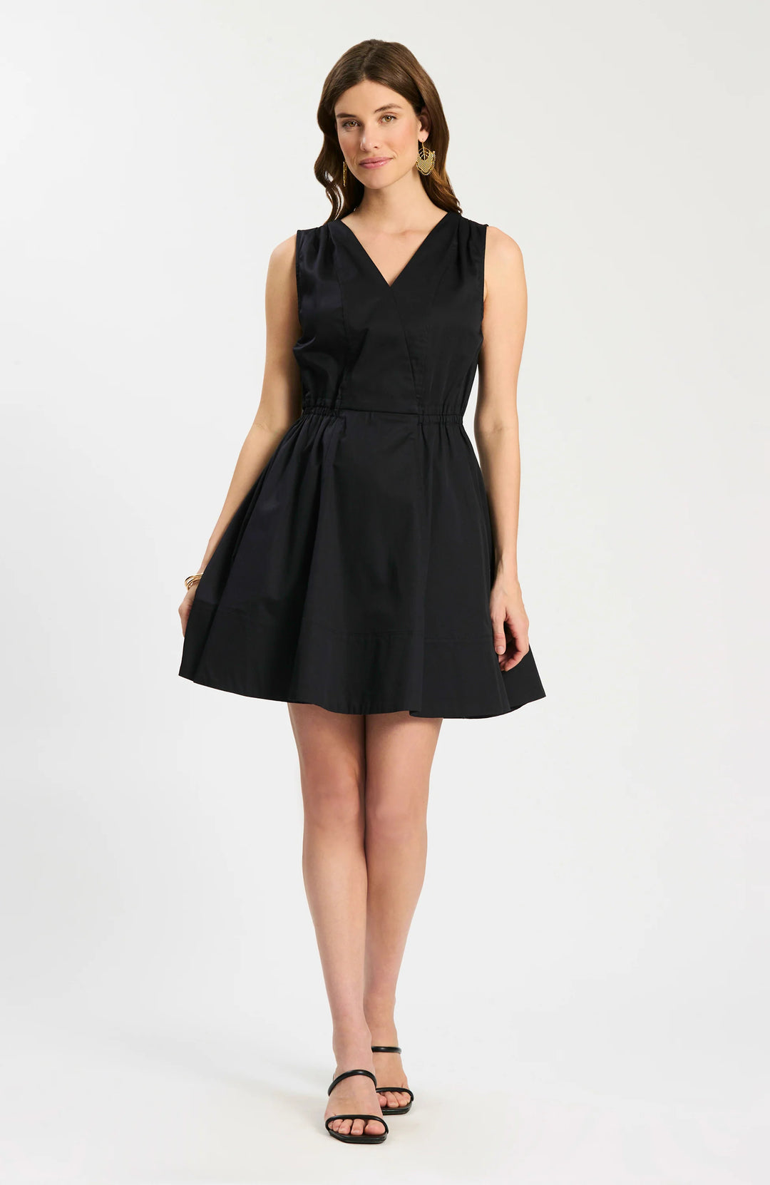 Model wearing Tyler Boe Emory Sleeveless Wrap Dress in black, front view belted with woven rattan belt, styled with black block-heel sandals and gold earrings, on white background.